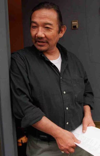 A color photograph of Kiyoshi leaning against a doorframe holding papers in his hands, a slight smile on his face as he seems to be mid-conversation