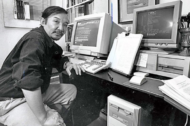 A photograph of Kiyoshi sitting at a desk surrounded by computer monitors and papers