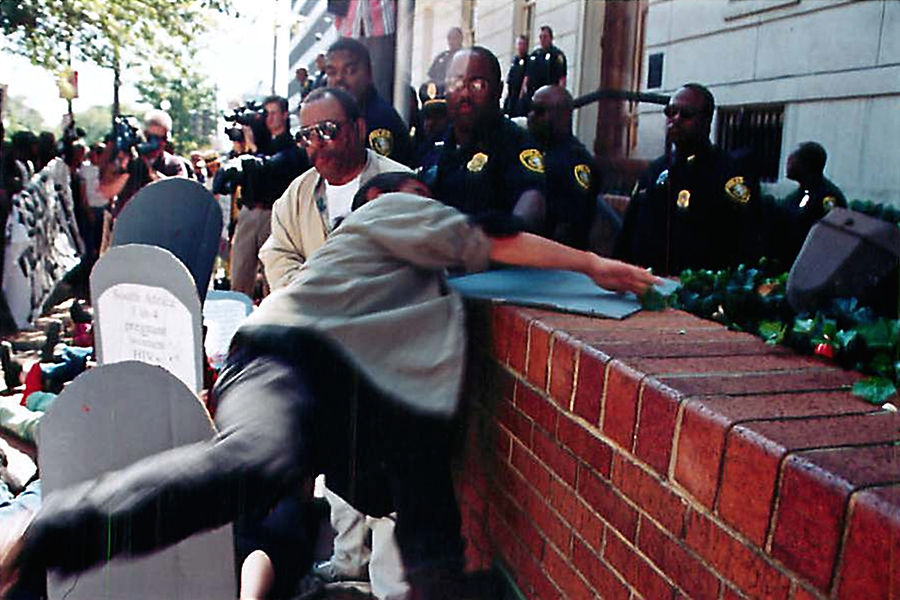 A color photo of Kiyoshi flinging himself over a low brick wall, with police officers and protestors surrounding him.