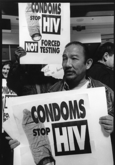A black and white photograph of Kiyoshi at a protest, holding a sign that reads "Condoms stop HIV, not forced testing."