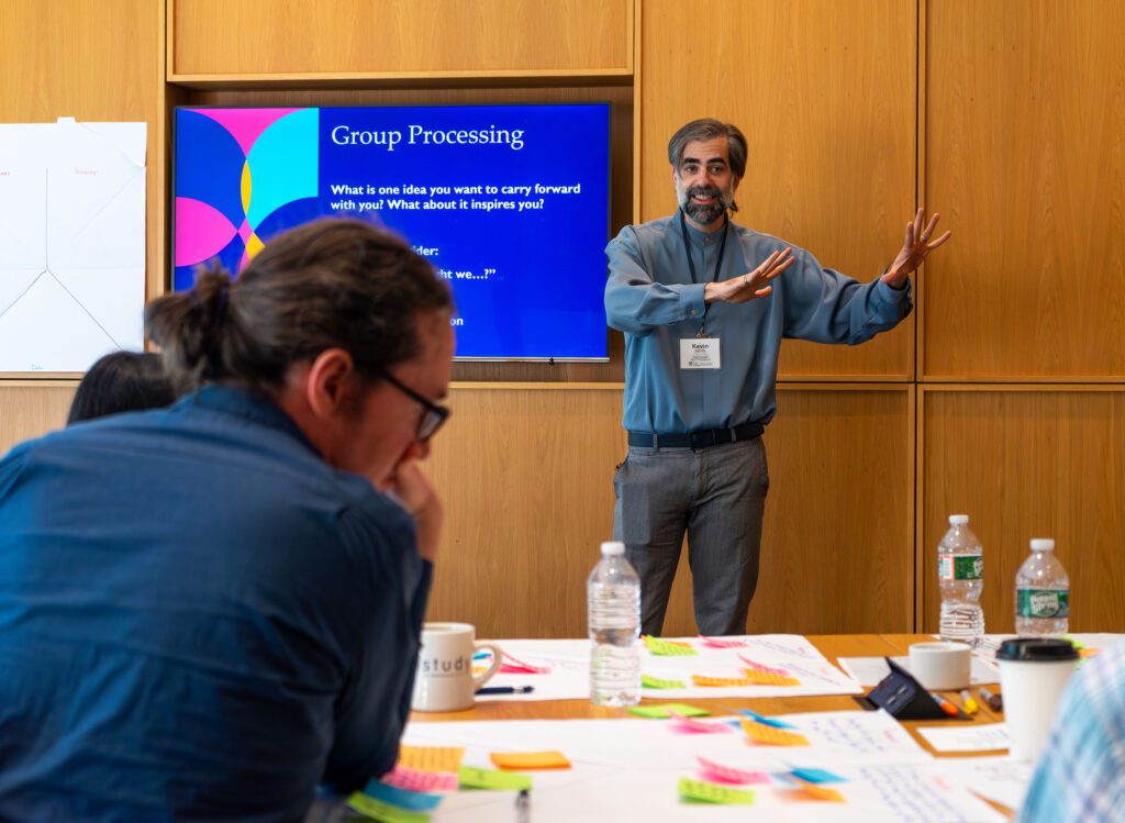 A photo of Eidos Director of Engagement Kevin Schott standing at the front of a room leading an activity while participants in the foreground look at papers on the table in front of them full of sticky notes.