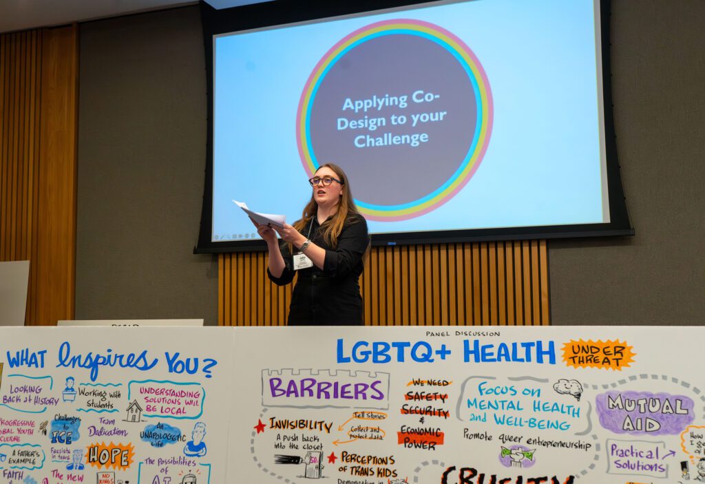 A photo of Julia Votto, Eidos' Associate Director of Innovative Projects, standing at the front of a room leading a discussion, with a screen behind her that reads "applying co-design to your challenge," and colorful boards full of visual notes in front of her.