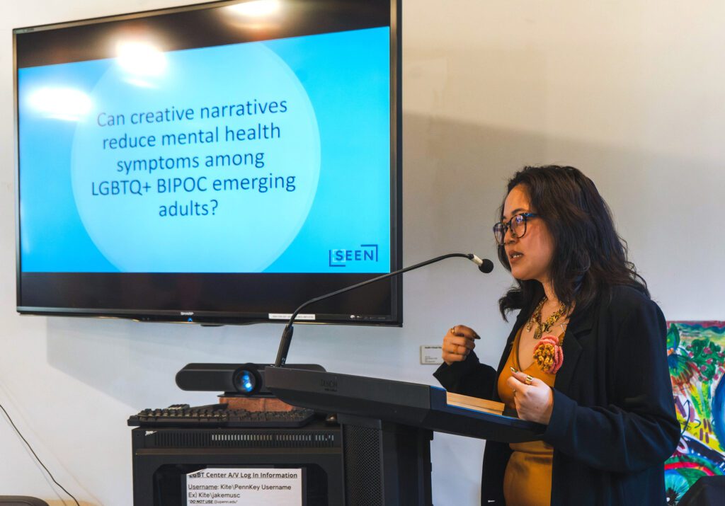 Dr. Jennifer Tran speaks at a podium with a screen behind her that reads "Can creative narratives reduce mental health symptoms among LGBTQ+ BIPOC emerging adults?"