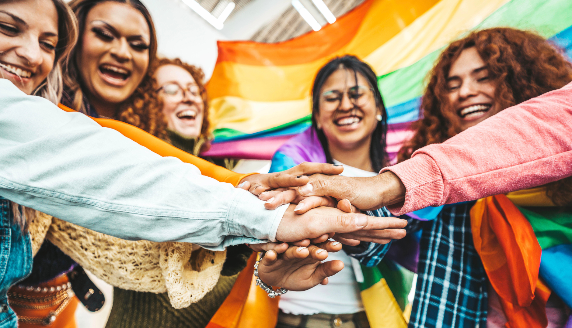 Photograph of diverse people smiling and putting their hands together, surrounded by colorful clothing and a pride flag behind them.