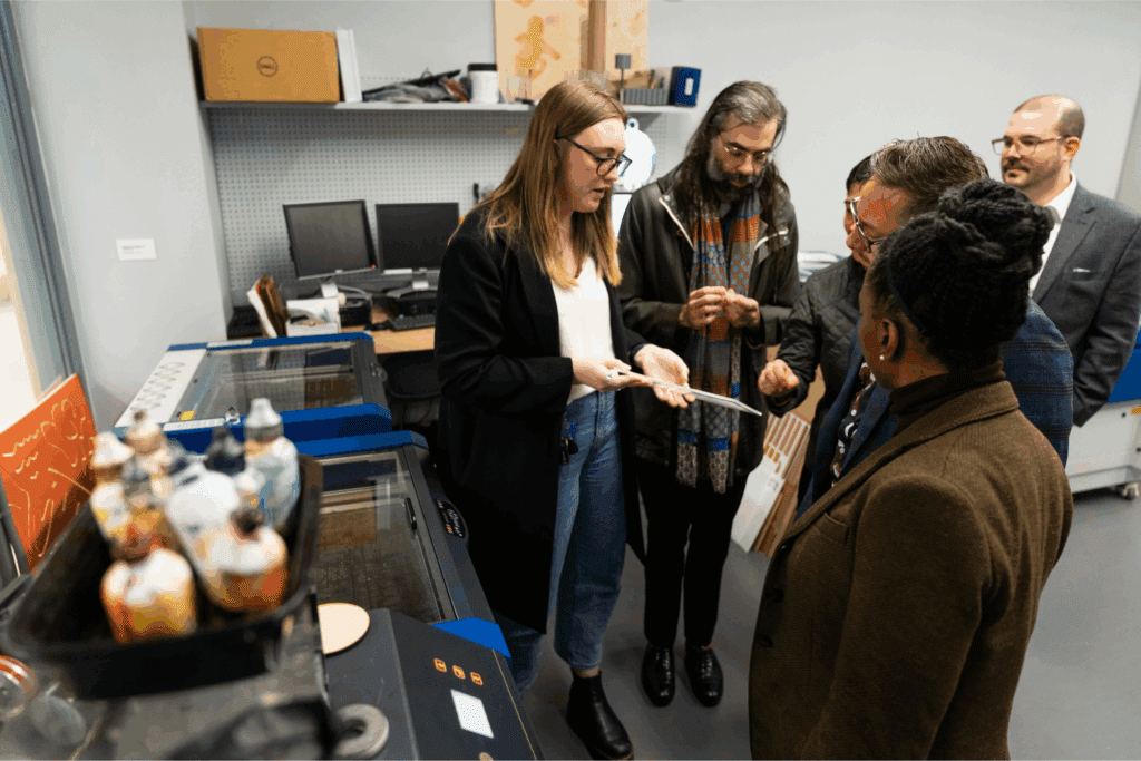 Photo of a group of people from the Eidos team gathered around an unintelligible object being demonstrated as part of a tour of a busy "makers space"