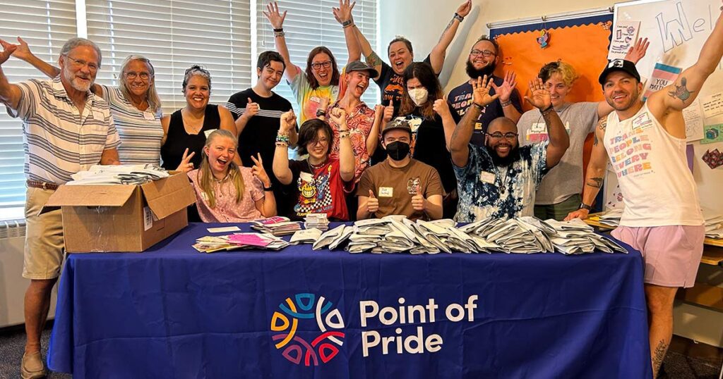 Photo of Point of Pride founders, volunteers, and others gathered together in joyous celebration at a table with a Point of Pride logo on the front