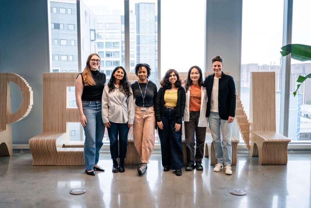 A group photograph of 6 people standing in front of a wooden PENN sign, smiling at the camera. On the left is Eidos Associate Director of Innovative Projects, Julia Votto and on the far right is entrepreneur Ashlie Grilz. Between them are 4 Penn students.