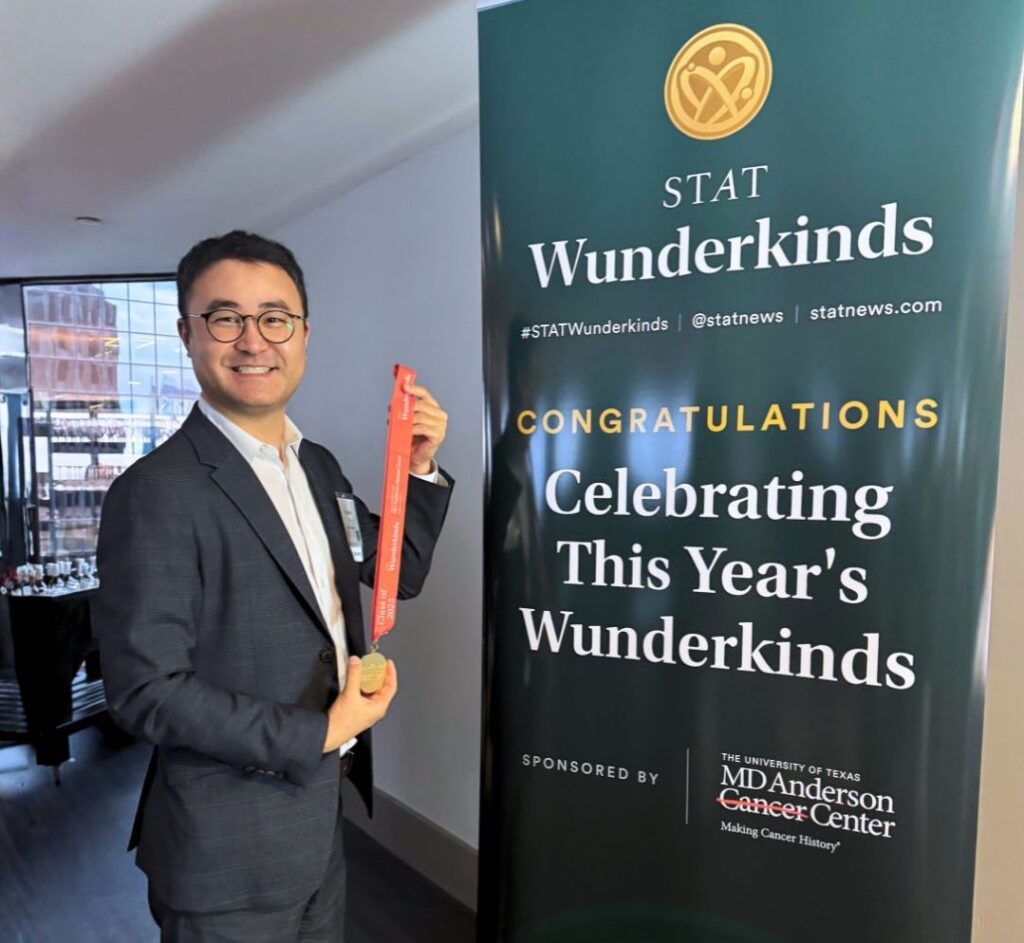 Photo of David Yu smiling and holding a medal next to a sign that reads "STAT Wunderkinds. Congratulations! Celebrating this year's wunderkinds"