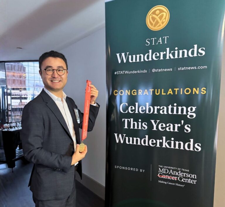 Photo of David Yu smiling and holding a medal next to a sign that reads "STAT Wunderkinds. Congratulations! Celebrating this year's wunderkinds"