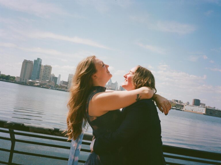 A photograph of Julia Votto and her partner Melanie smiling with their arms around each other, the Delaware River in the background