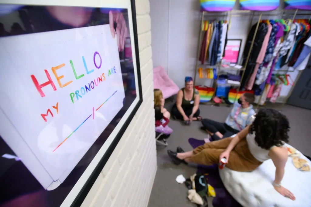 A photograph of a group of people hanging out in an LGBTQ+ center, with a sign on the left that says "my pronouns are..."