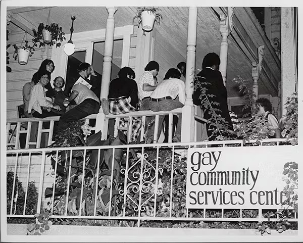 an old black and white photograph of a porch full of people gathering, with a sign on the front that says "gay community services center"