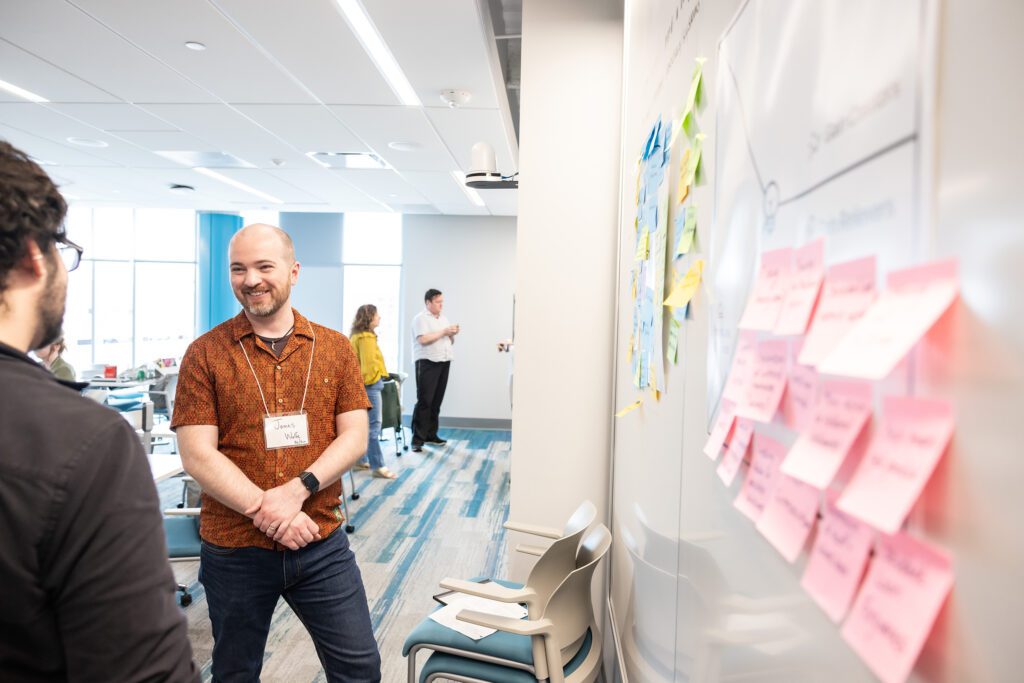 A photo of James Wolfe speaking with someone with a smile on his face, with a whiteboard to his right covered in sticky notes.