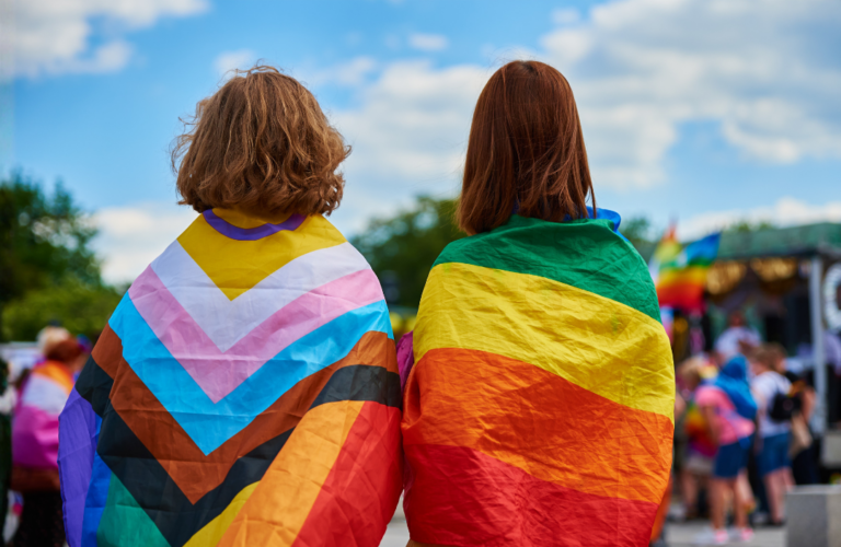 A photo of two people standing side by side facing away from the camera, cloaked in two pride flags