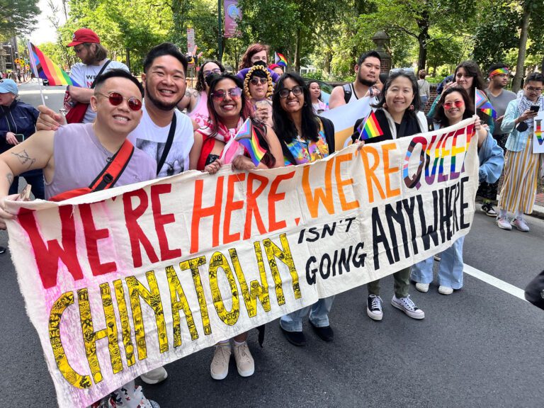 A photo of a group of asian americans wearing rainbow pride outfits and accessories, holding a large banner that reads "We're here, we're queer, chinatown isn't going anywhere."
