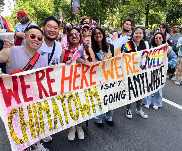 A photo of a group of asian americans wearing rainbow pride outfits and accessories, holding a large banner that reads "We're here, we're queer, chinatown isn't going anywhere."